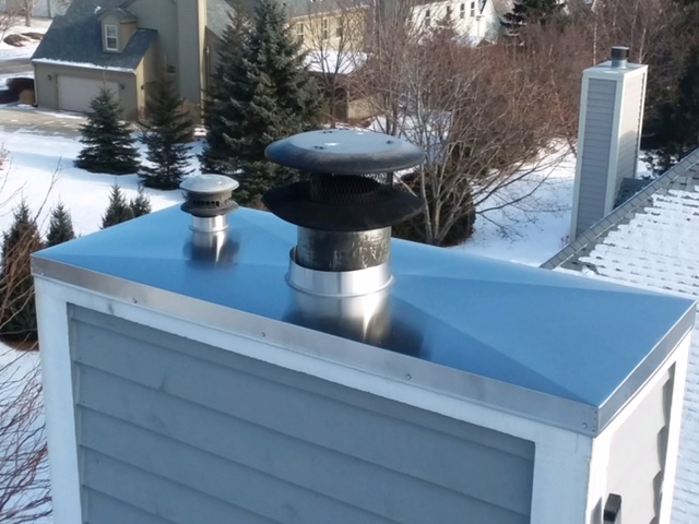 Blue siding chimney with white trim featuring a newly installed stainless steel chase cover with snow in the background